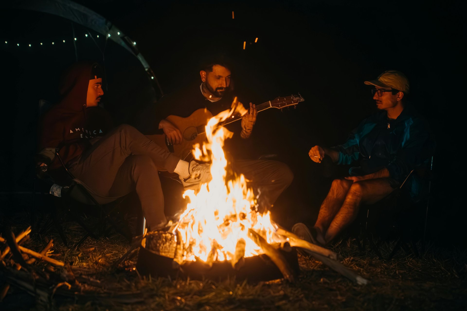 Three guests around a bonfire at Windmill Inn — one playing guitar, fire at the centre, string lights overhead