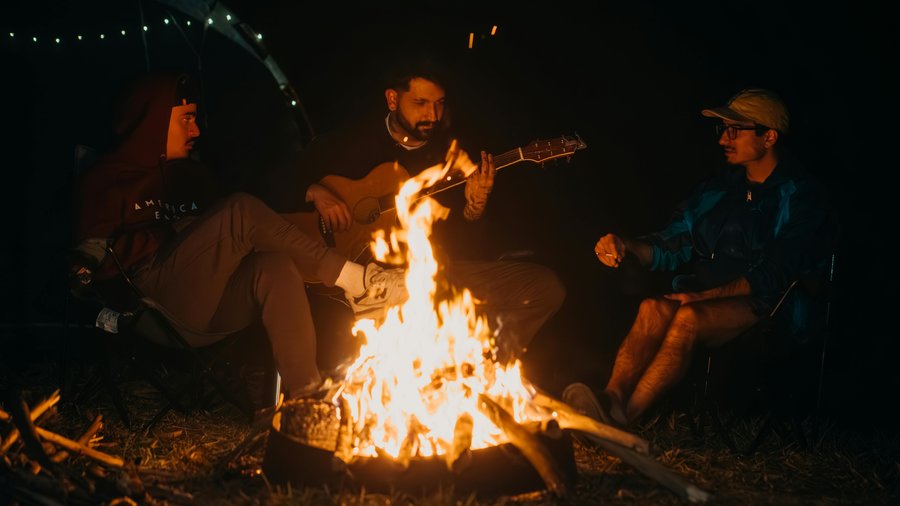 Three guests around a bonfire at night — one playing guitar, fire warming the circle