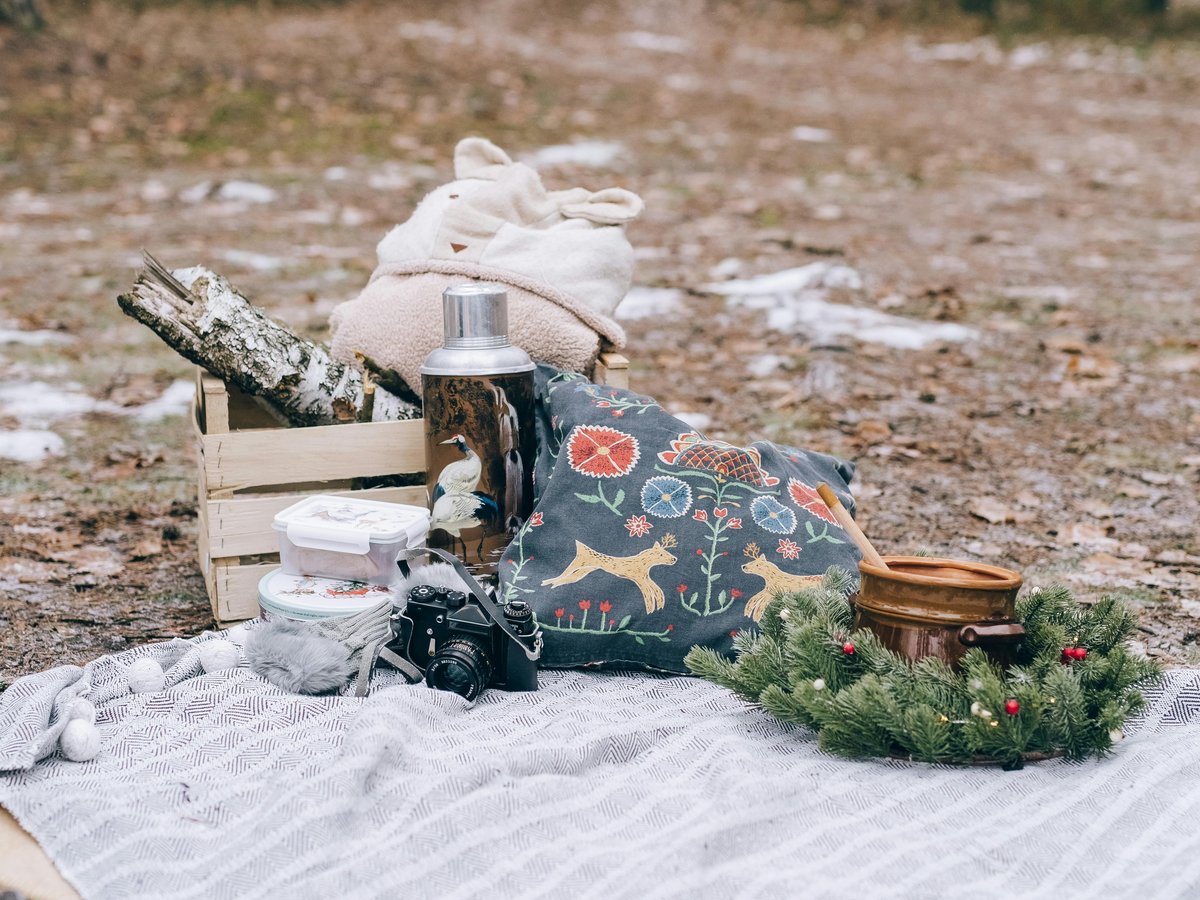 A winter forest picnic setup — thermos, embroidered cushion, and a small crate of firewood on a blanket