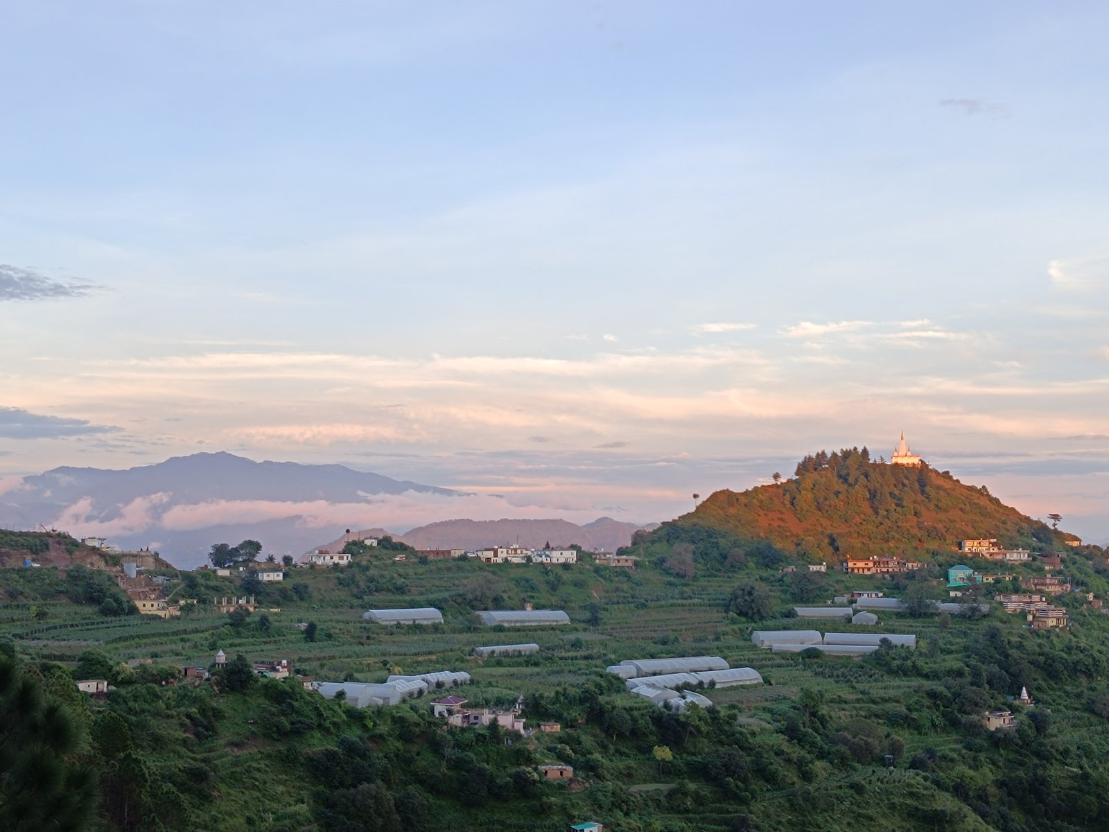 Sunset view over the village near Windmill Inn — terraced farms, polytunnels, and a hilltop temple with layered Himachal ranges in the distance