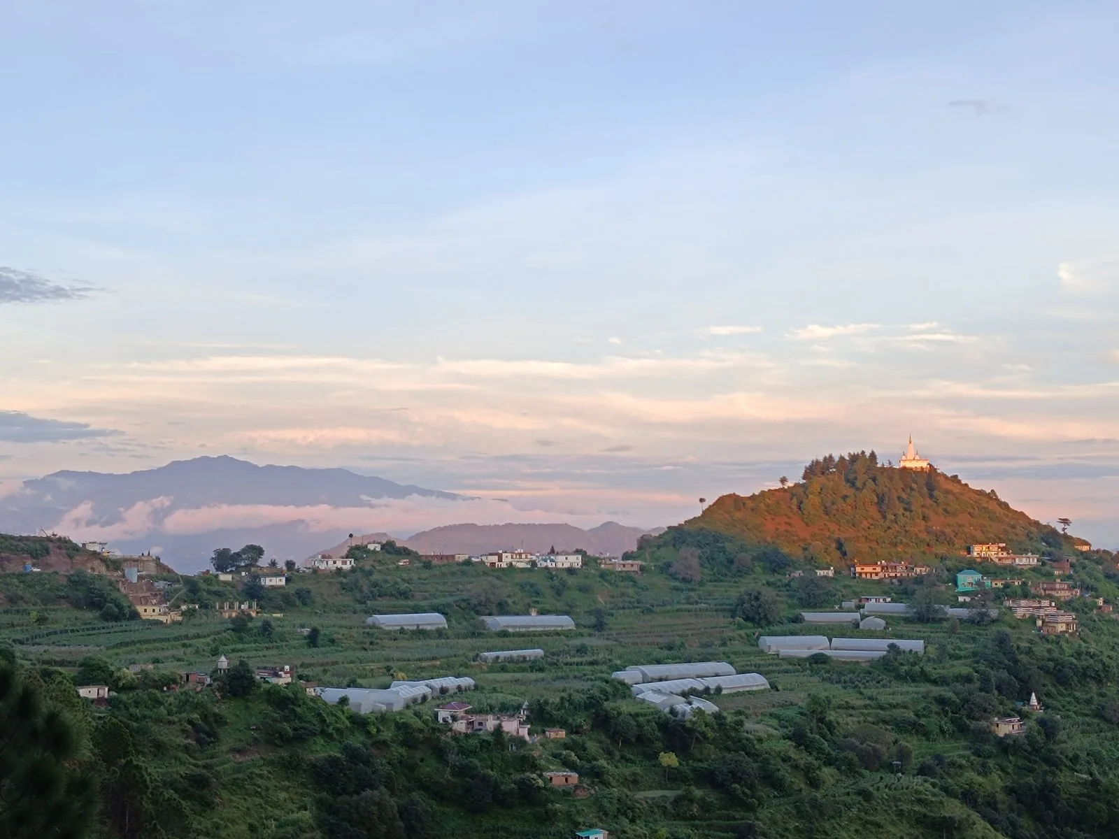 Sunset view over the village near Windmill Inn — terraced farms, polytunnels, and a hilltop temple with layered Himachal ranges in the distance