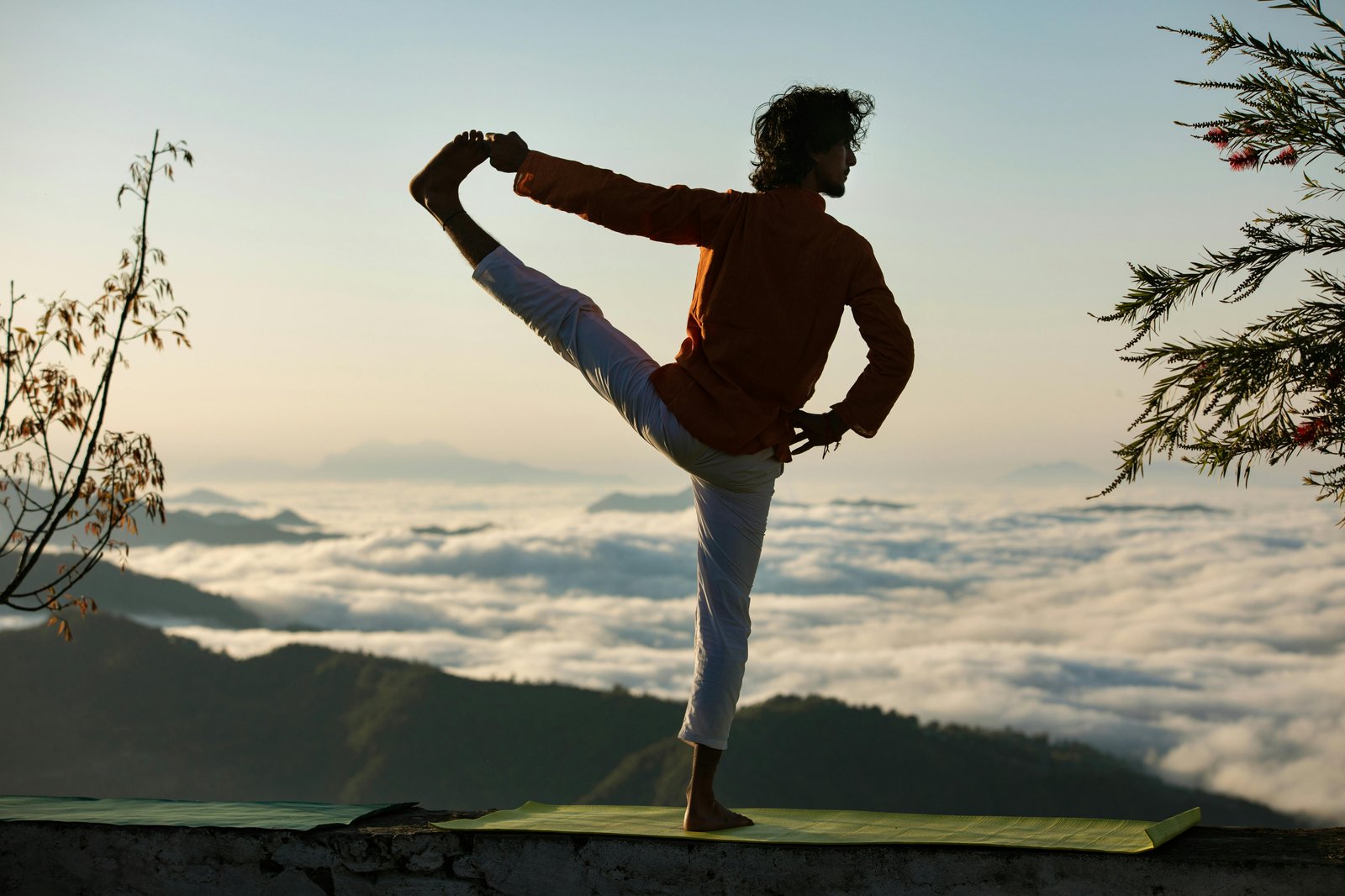A yoga practitioner in dancer's pose on a mat at sunrise, with a sea of clouds and mountain ridges in the distance