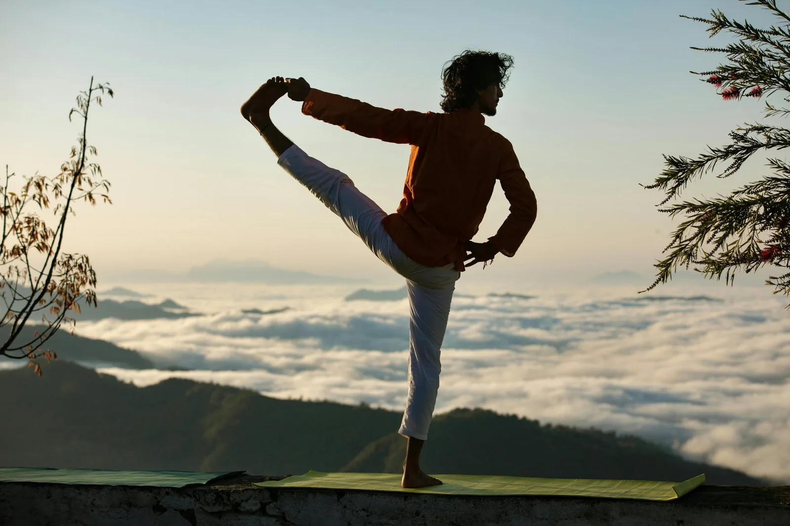 A yoga practitioner in dancer's pose on a mat at sunrise, with a sea of clouds and mountain ridges in the distance